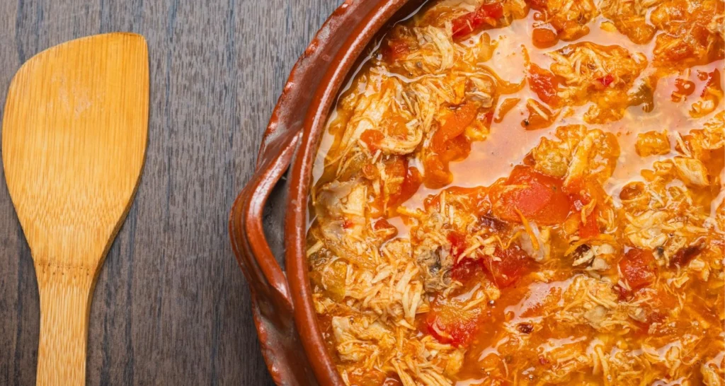 A flavorful shredded chicken dish cooked with tomatoes and spices in a clay pot, set on a wooden table with a wooden spatula beside it.