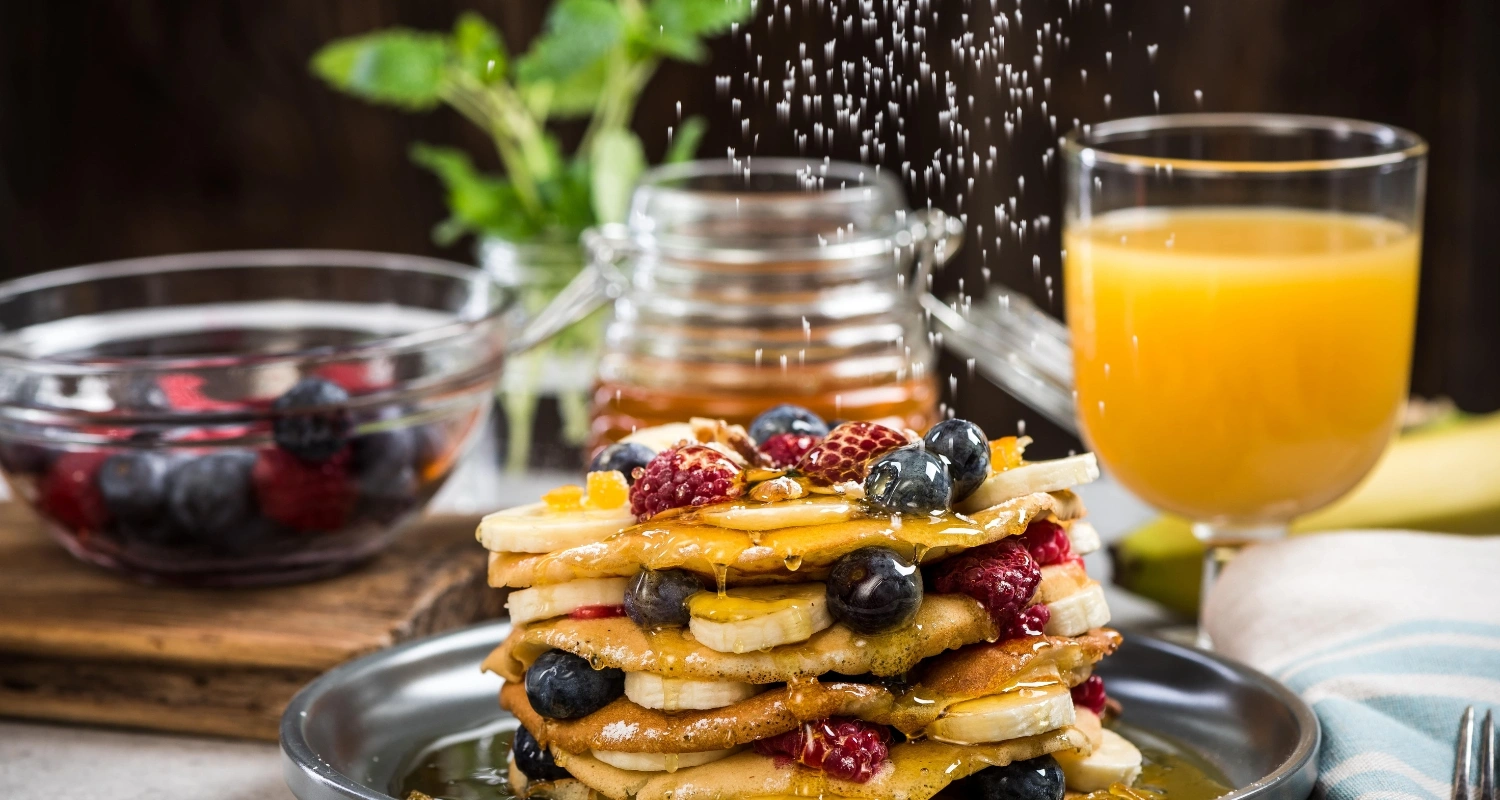 A stack of golden pancakes topped with fresh berries, banana slices, and drizzled with syrup, dusted with powdered sugar, alongside a glass of orange juice and a bowl of mixed fruit.