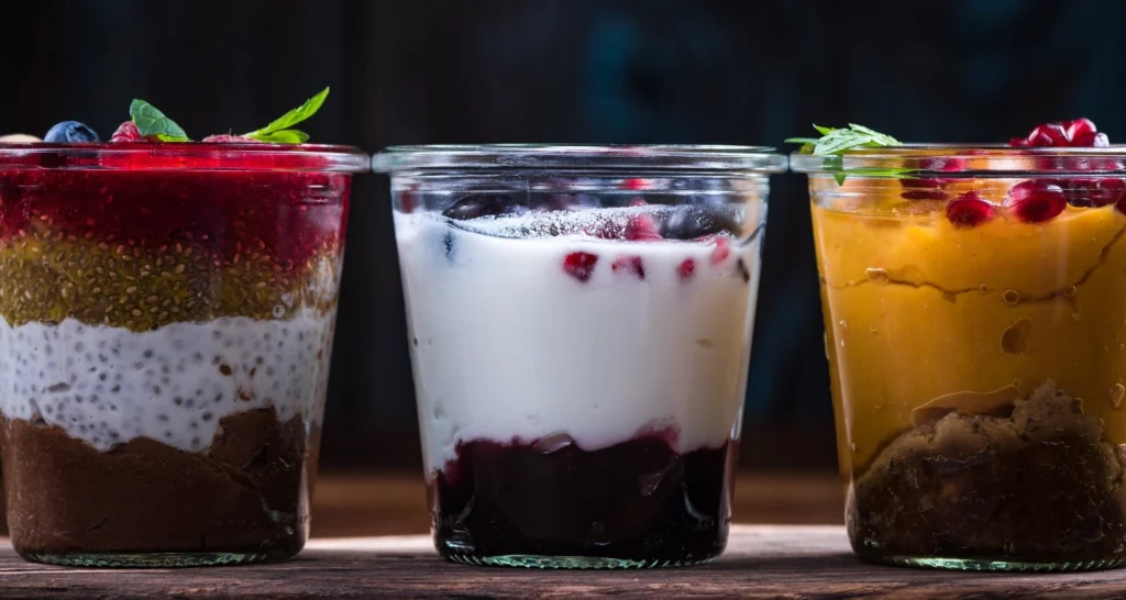 Three vibrant dessert jars featuring layers of colorful ingredients: one with chia pudding, chocolate, and raspberry compote; one with yogurt, berries, and pomegranate seeds; and one with mango puree, pomegranate seeds, and a spiced crumble base.