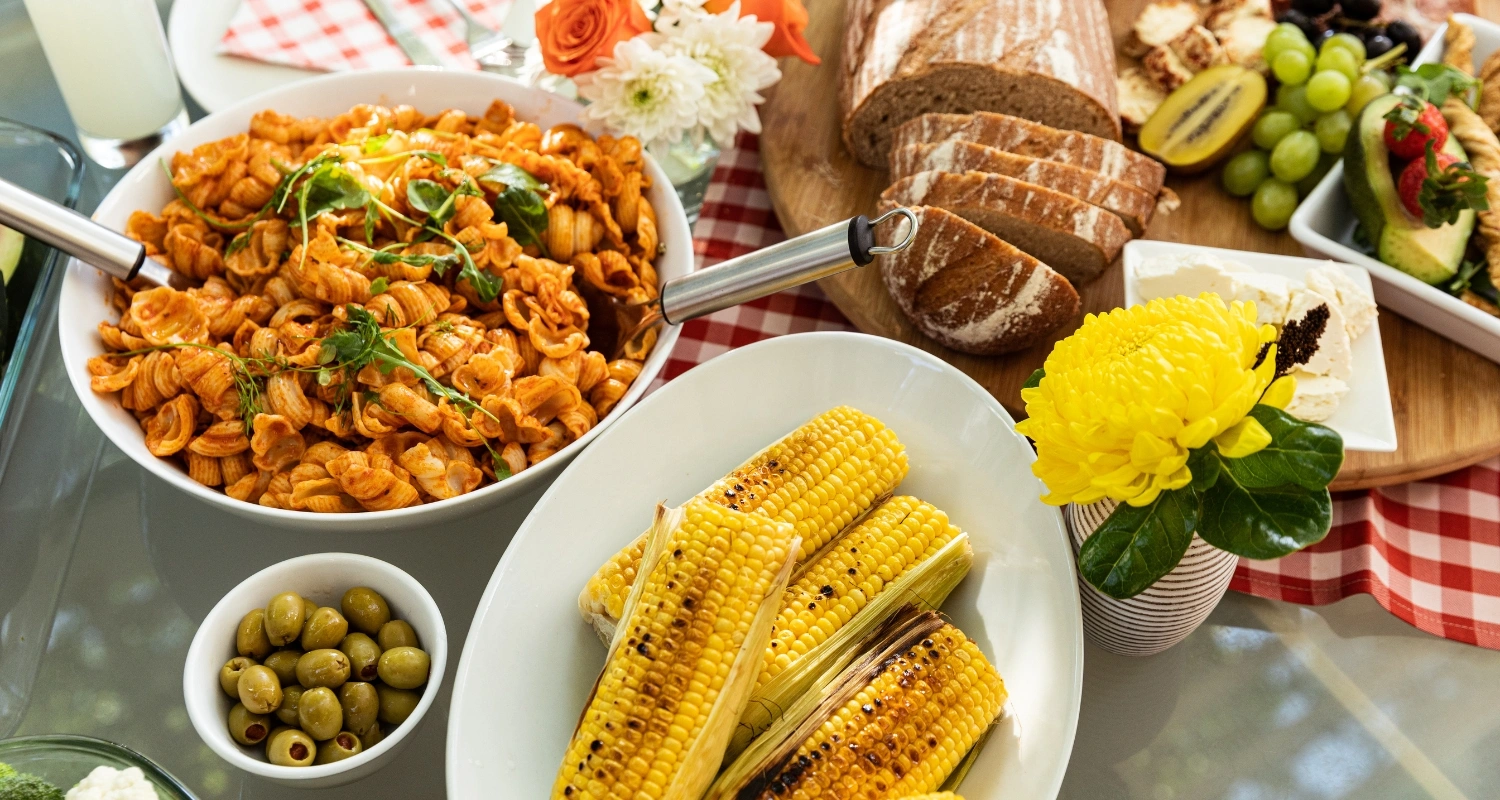 A festive Puerto Rican spread with pasta, grilled corn, fresh bread, olives, fruits, and cheeses on a checkered table, accented with tropical flowers.