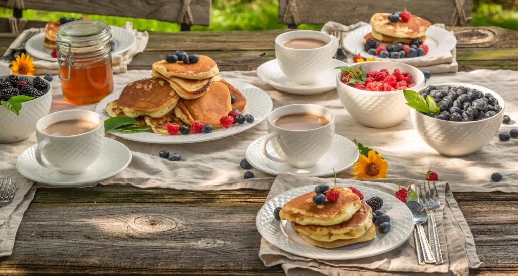 A rustic outdoor table set for a CMS Pancake Breakfast, featuring stacks of pancakes with fresh berries, bowls of fruit, cups of coffee, and a jar of honey.

