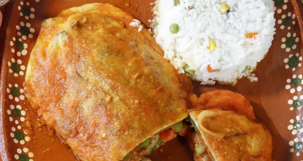 A traditional Puerto Rican dish featuring stuffed peppers in a rich tomato-based sauce, served alongside white rice with mixed vegetables, presented on a rustic ceramic plate.