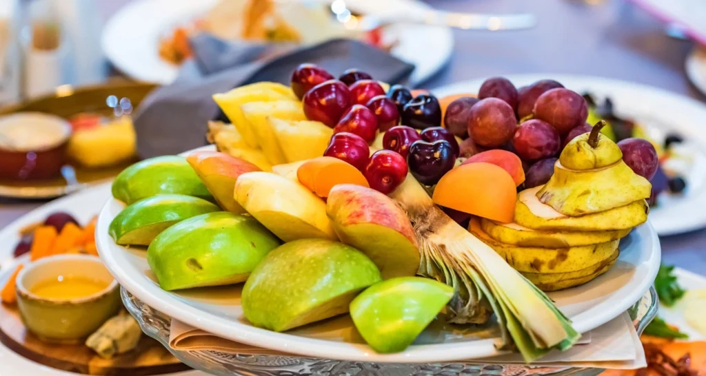 A beautifully arranged platter of fresh fruits, including green apples, bananas, pineapple, grapes, cherries, apricots, and pears.