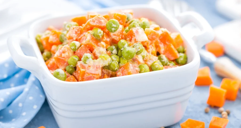 A creamy dish of peas and diced carrots served in a white ceramic bowl with handles, placed on a blue tablecloth with scattered carrot pieces.