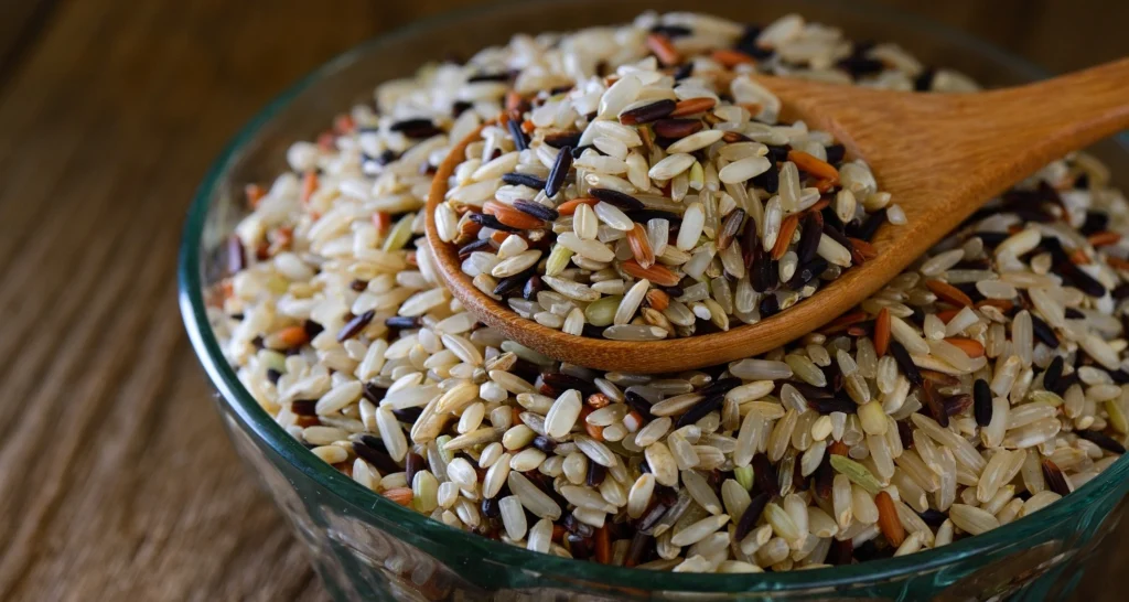 A close-up of a glass bowl filled with a colorful wild rice blend, including black, red, and brown grains, with a wooden spoon scooping the rice.