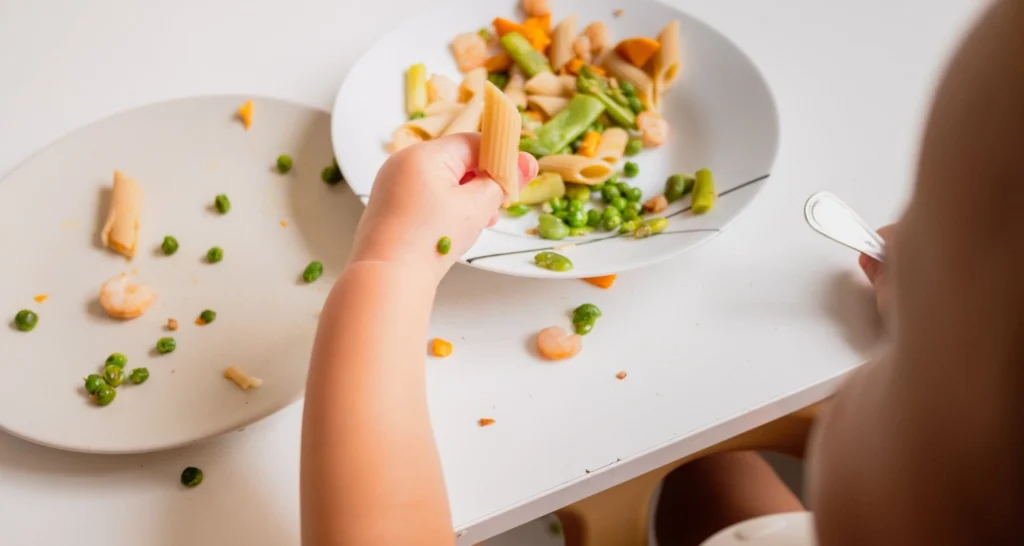 A toddler enjoying a healthy lunch of colorful pasta with peas, shrimp, and vegetables, using their hands to self-feed at the dining table.