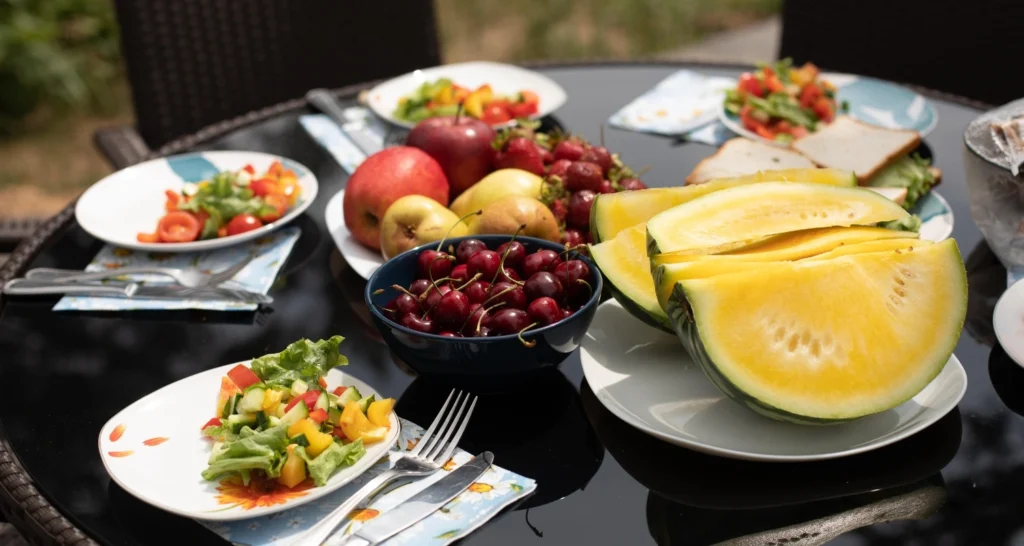  summer outdoor lunch table with plates of fresh salad, yellow watermelon slices, cherries, apples, pears, and sandwiches.